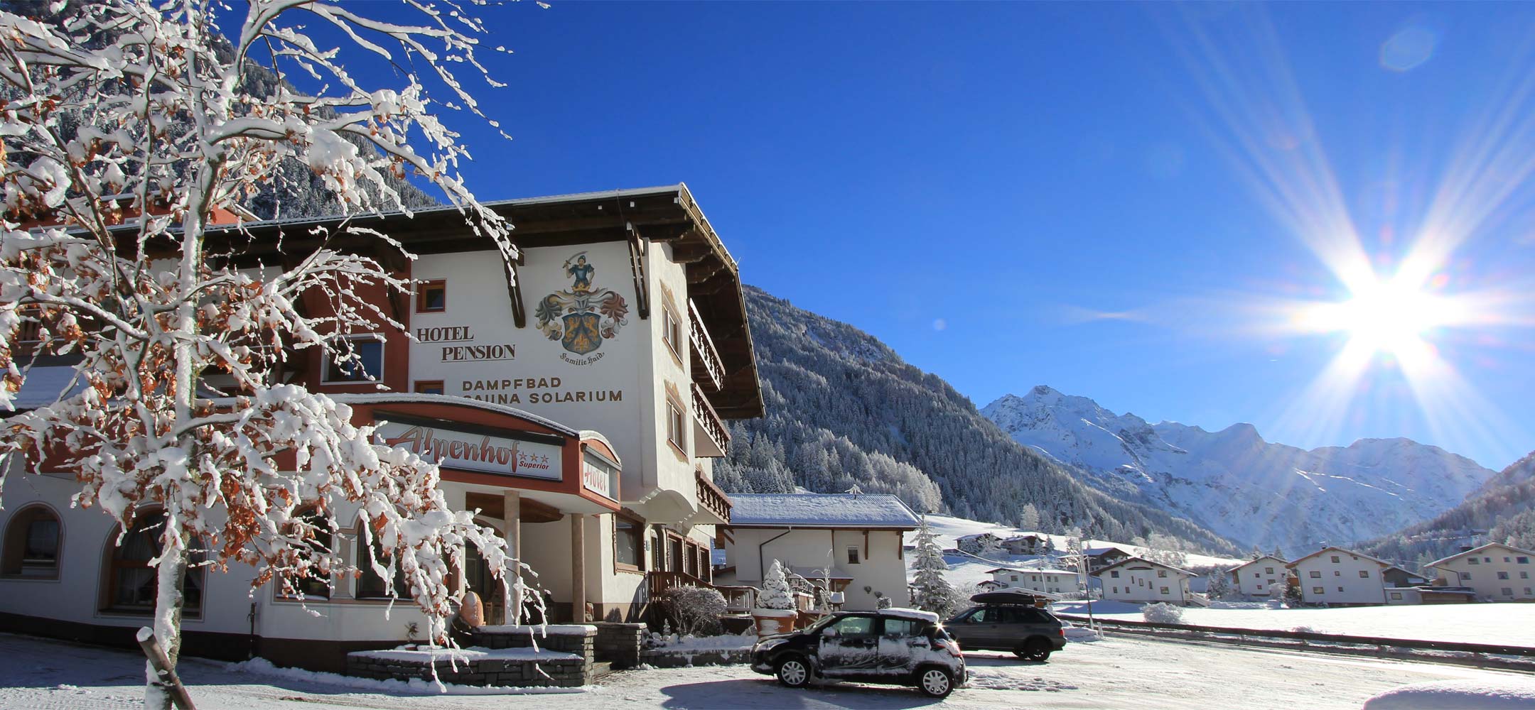 Hotel Alpenhof Pitztal bei Sonnenschein im Schnee, vor schneebedeckten Bergen mit klarblauem Himmel und glitzerndem Sonnenlicht.
