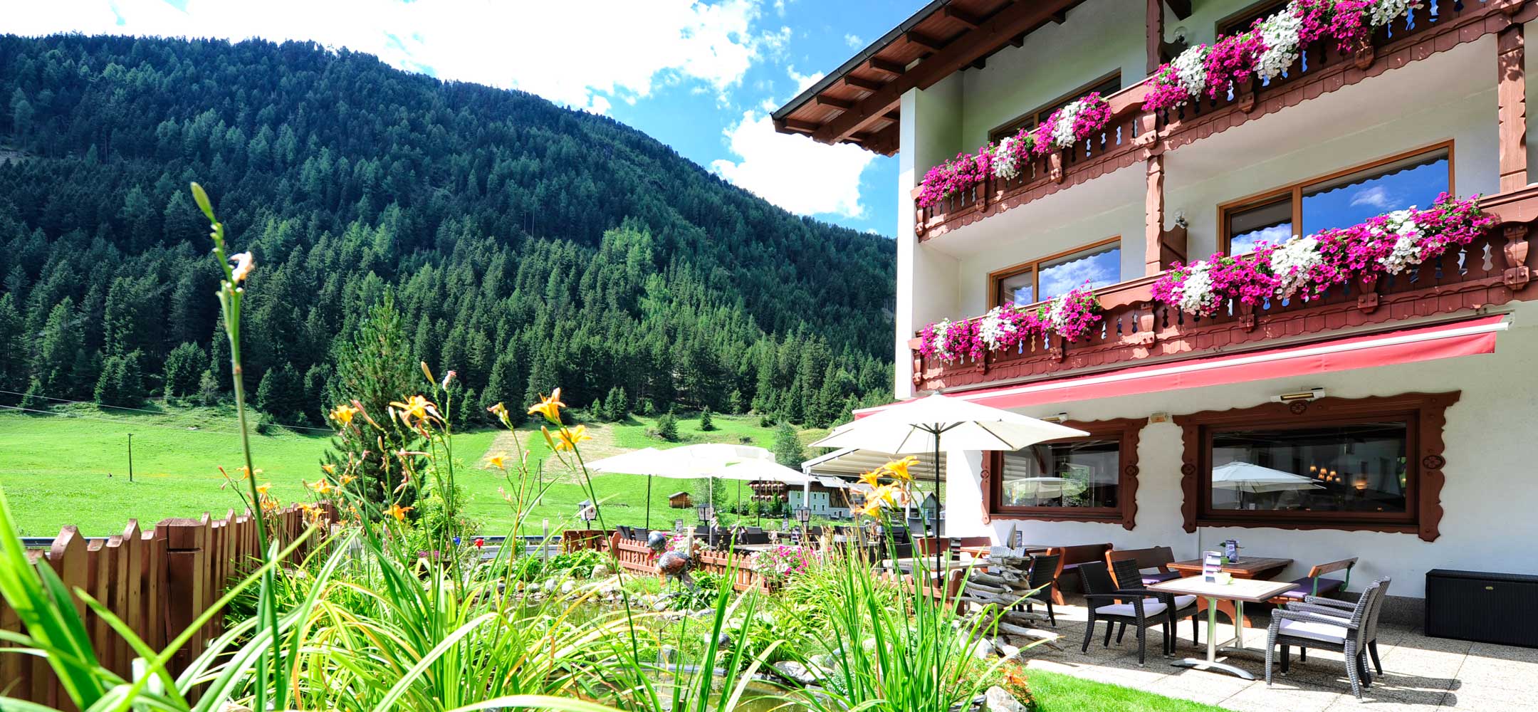 Sonnige Terrasse des Hotels Alpenhof, umgeben von Blumenbeeten und grünen Wiesen.