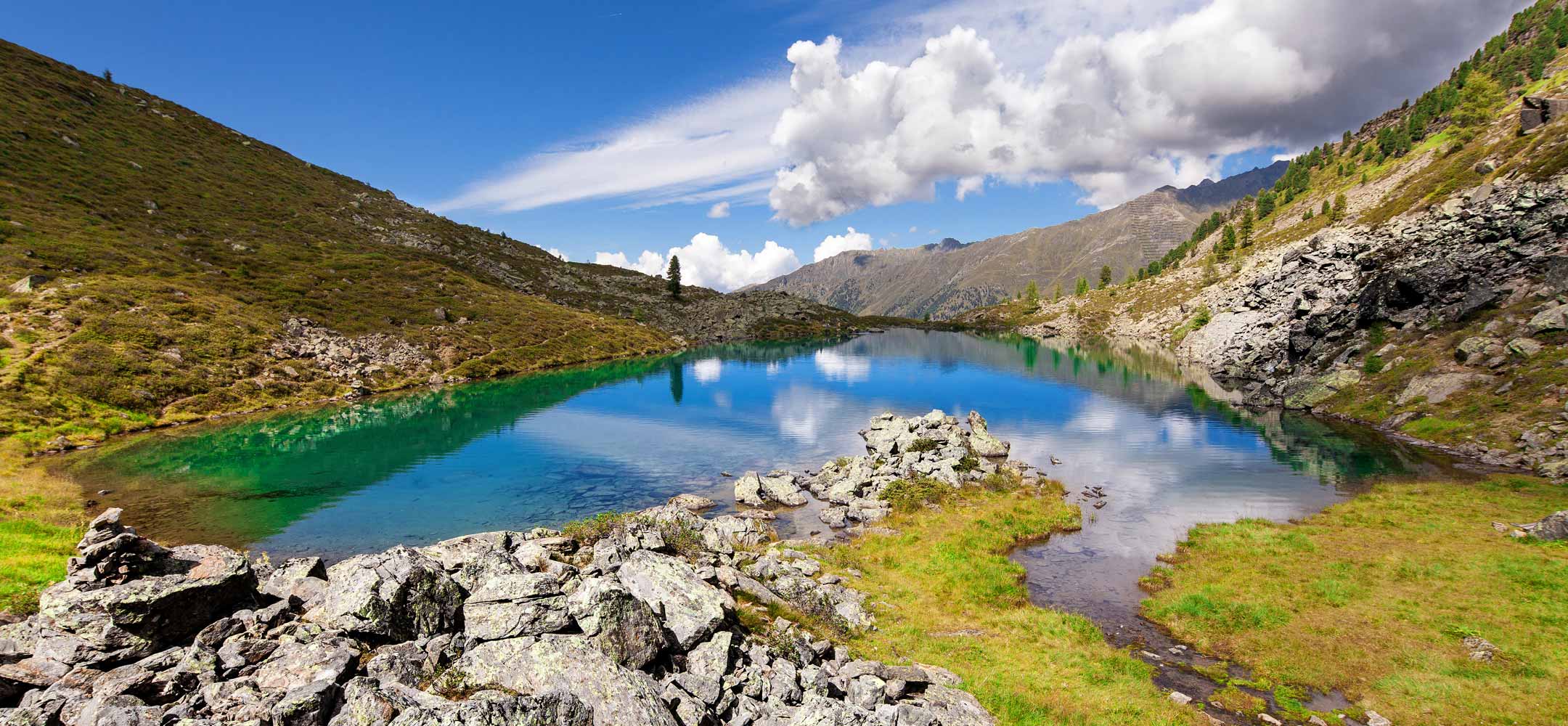 Kristallklarer Bergsee mit Felsufer, umgeben von grünen Hängen und blauen Himmel.
