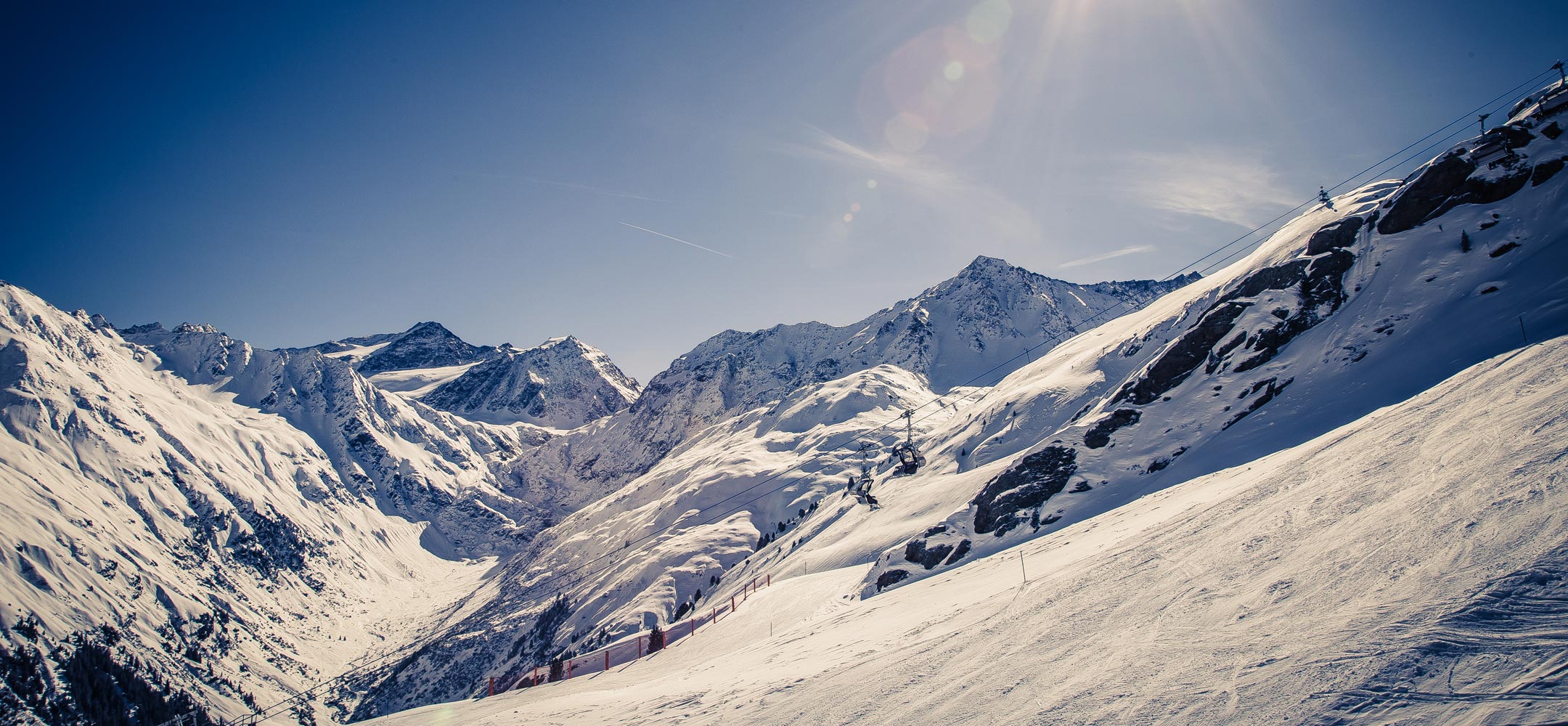 Verschneite Alpenlandschaft mit sonnigem Himmel, Skipiste und Sessellift in hochalpiner Umgebung.